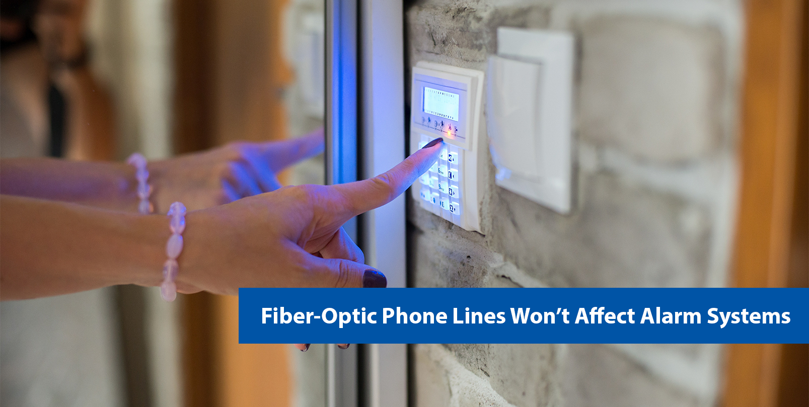 Photo shows a person's hand pressing a button on a wall-mounted alarm system. Text on image says "Fiber-Optic Phone Lines Won’t Affect Alarm Systems."