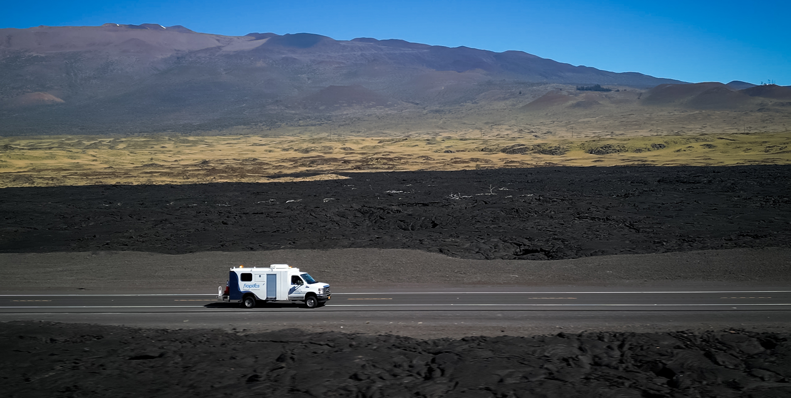 Fioptics van driving across road with remnants of a lava field in foreground and mountain the background.